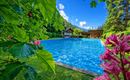A beautiful swimming pool surrounded by trees and colorful flowers. In the background, cozy wooden houses and a clear blue sky can be seen.