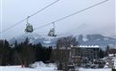 A snowy landscape with ski lifts in the background. Densely wooded mountains are visible in the distance.