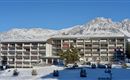 A modern hotel building, surrounded by snow-capped mountains and a clear blue sky. Winter is clearly visible with a white blanket of snow and green fir houses in the foreground.