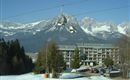 A large mountain landscape with snow-covered peaks and a ski lift in the foreground. In the background, a hotel and a parking facility are visible.