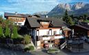 Ein malerisches Haus in den Alpen mit traditionellen Holzverkleidungen. Im Hintergrund sind majestätische Berge und ein klarer Himmel zu sehen.
