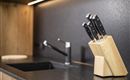 A modern kitchen environment with a wooden cutting block holding five sharp knives. In the background, a sink and a faucet are visible.
