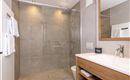 A modern bathroom with a large shower and gray tiles. The wash area has a wooden cabinet and a mirror.