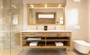 A modern bathroom with a large mirror wall and an elegant wooden washstand. The shower is made of glass and there are fresh towels on the open shelf.