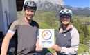 A pair of cyclists stands next to their bicycles holding a sign with the logo "bett+bike". In the background, mountains and a clear blue sky can be seen.