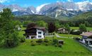 A picturesque landscape with a traditional house and majestic mountains in the background.  
Surrounded by green meadows and trees, the scene radiates tranquility and natural beauty.