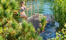 A woman in a swimsuit stands on a steps leading to a body of water. In the background, tall plants and a pier can be seen.