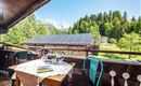 Een gezellige balkon met een tafel en stoelen, omringd door bomen en bergen. Op de achtergrond zijn zonnepanelen te zien, onder een heldere blauwe lucht.