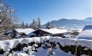 A winter landscape with snow-covered roofs and trees. In the background, gentle mountains and a clear blue sky can be seen.
