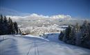 Eine schneebedeckte Landschaft mit Bergen im Hintergrund und einem kleinen Dorf im Tal. Der Himmel ist klar und blau, was eine friedliche Winteratmosphäre schafft.