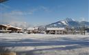 A winter landscape with snow-covered huts and mountains in the background. The sky is clear and blue.