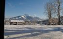 A snowy landscape with an impressive mountain in the background. In the foreground, there is a house and there are sporadic trees.