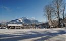 A winter landscape with snow-covered fields and a view of snow-covered mountains. In the foreground, there is a cozy house under a clear blue sky.