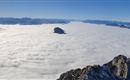 An impressive mountain landscape above a thick layer of clouds. In the background, snow-covered peaks and a clear blue sky can be seen.