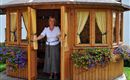 A woman stands in the open door of a cozy wooden house. Flowers adorn the windows, creating an inviting atmosphere.