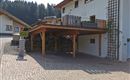 Ein modernes Haus mit einer überdachten Terrasse und einem gepflasterten Parkplatz. Im Hintergrund sind grüne Bäume und eine ruhige Landschaft sichtbar.