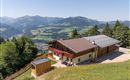 Ein modernes Holzhaus auf einer Anhöhe mit Blick auf die umliegenden Berge. Die Landschaft ist grün und hügelig, unter einem klaren blauen Himmel.