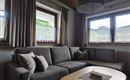 A cozy living room with a gray sofa and a wooden table. The windows are adorned with curtains and provide a view of the outdoors.