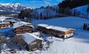 A picturesque snowy landscape with several wooden houses in the foreground. In the background, the snow-covered mountains and a blue sky can be seen.