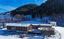 A rustic hotel in a snow-covered landscape. In the background, forested mountains and a blue sky can be seen.