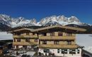A cozy chalet in the mountains with snow-covered peaks in the background. Clear blue skies and a tranquil setting.
