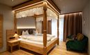 A cozy bedroom with a wooden four-poster bed. There is a small table and a green couch, as well as large windows with curtains.