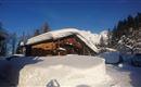 A snowy landscape with a traditional wooden house and mountains in the background. The sky is clear and blue, creating a calm winter atmosphere.