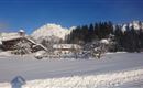 A winter landscape with snow-covered fields and mountains in the background. In the foreground stands a traditional wooden house surrounded by snow-covered trees.