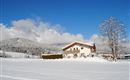 A snow-covered landscape with a modern house in the foreground. In the background, the snow-capped mountains rise under a blue sky.