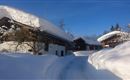 A snowy landscape with traditional wooden houses. The sky is clear and blue, and the path is clearly visible.
