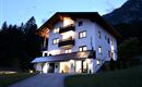 Ein modernes Haus im Alpenstil, das abends beleuchtet ist. Umgeben von Bäumen und Bergen, strahlt es eine einladende Atmosphäre aus.