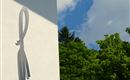 Ein modernes Gebäudewand mit einem stilisierten Logo. Im Hintergrund sind grüne Bäume und ein blauer Himmel mit einigen Wolken zu sehen.