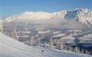 Eine verschneite Berglandschaft mit übersähten Bäumen. Ein Skifahrer fährt die Piste hinunter, während die Berge im Hintergrund majestätisch in den Himmel ragen.