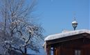 A snow-covered roof of a wooden house stands against a clear, blue sky. In the background, a tree with snow-white branches can be seen.