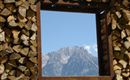 Ein Fensterrahmen aus Holzstämmen zeigt einen Blick auf einen majestätischen Berg und den blauen Himmel. Die Szene vermittelt Ruhe und Naturnähe.