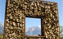 Een houten frame met een rechthoekige uitsnede, gevuld met houtblokken. Op de achtergrond zijn bergen en een heldere blauwe lucht te zien.