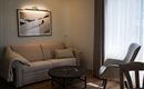 Een gezellige woonkamer met een lichte bank en een moderne fauteuil. Een ronde tafel en een groot raam zorgen voor veel daglicht.
