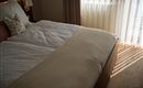 Een gezellige slaapkamer met een groot bed en zachte dekens. Daglicht stroomt door de gordijnen en werpt interessante schaduwen op de vloer.