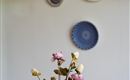Een elegante kan met kleurrijke bloemen staat op een tafel. Op de achtergrond hangen decoratieve borden aan de muur.