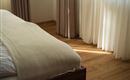Een gezellige slaapkamer met een bed en een lichte, neutrale deken. Zachte gordijnen laten warm licht binnen.
