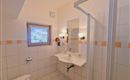 A bright bathroom with a shower, sink, and mirror. It has white tiles and a window that lets in light.