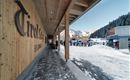 A modern entrance area with wood paneling in a snow-covered environment. In the background, people can be seen preparing for a ski trip.