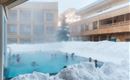 A pool in the middle of snow with several people relaxing in the water. In the background, the wooden buildings of an accommodation can be seen.