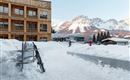 A picturesque winter resort with snow-covered mountains in the background. In the foreground, there is ski equipment and people walking in the snow.