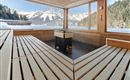 A cozy sauna area with wooden benches and a fireplace. In the background, snow-covered mountains stretch under a blue sky.