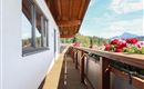 Ein heller Balkon mit Holzfußboden und Blumenkästen. Im Hintergrund sind Berge und ein blauer Himmel zu sehen.