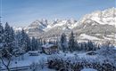 A snowy winter landscape with majestic mountains in the background. In the foreground stands a charming wooden house surrounded by snow-covered trees.