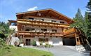 A lovely chalet in alpine style with wooden balconies adorned with carvings and beautiful flowers. Surrounded by trees and a green meadow.