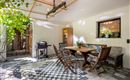 A cozy terrace with a dining table and wooden chairs. In the background, there is a grill and a plant providing shade.