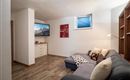 A cozy lounge with a dark blue sofa and a television on the wall. The room has wooden flooring and is brightly lit.